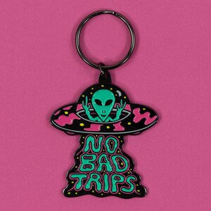 May include: Black metal keychain with a pink and black UFO design. The UFO has a green alien inside with its arms raised. The text "NO BAD TRIPS" is written in green and black below the UFO.