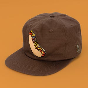 May include: A brown baseball cap with a textured grid pattern, featuring an embroidered graphic of a cartoon hot dog with a smiling face and yellow mustard on the front panel, and a small embroidered logo on the side of the cap.