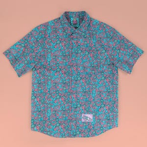 May include: Short-sleeved button-down shirt with a vibrant floral pattern in teal and pink. The shirt features a classic collar, a chest pocket, and a small logo patch near the hem. The shirt is laid flat on a peach-colored surface.