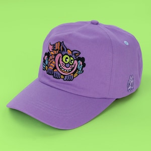 May include: A lavender baseball cap featuring an embroidered graphic of a grinning, orange and purple striped cat with large green eyes. The design includes small blue and black floral accents. A small purple rabbit character is embroidered on the side.