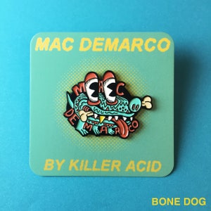 May include: A green, red, and yellow enamel pin featuring a cartoon dog with the text "MAC DEMARCO" and "BY KILLER ACID" on a blue background. The pin is labeled "BONE DOG".