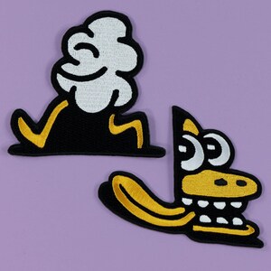 May include: Two embroidered patches on a purple background. One patch features a black shape with a white cloud-like top and yellow accents. The other patch is a yellow and black cartoon alligator head with white eyes.