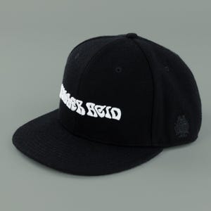 May include: Black baseball cap with a flat brim. The cap features white embroidered text and a small emblem on the side. The hat is made of a textured fabric and has a classic, structured design.