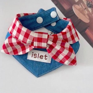 May include: A denim dog bandana with a red and white checkered bow. The bandana has a blue denim base with white snap closures. The word "islet" is printed on a white label.