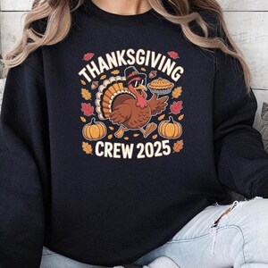 May include: Black sweatshirt with a Thanksgiving-themed graphic. The design features a turkey wearing sunglasses and a hat, holding a pie, with pumpkins and fall leaves. The text reads "Thanksgiving Crew 2025".