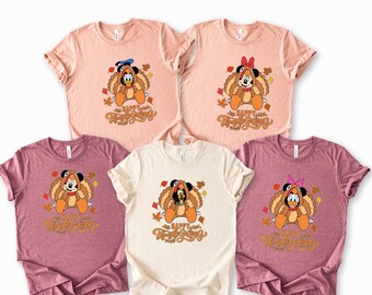 May include: Five short-sleeved t-shirts in various colors: light pink, burgundy, and cream. Each shirt features a cartoon character dressed as a turkey with the text "Happy Thanksgiving" in a script font. Fall leaves are scattered around the characters.