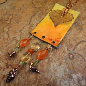 Puede incluir: Un collar de cobre con un colgante rectangular que presenta una hoja prensada y un degradado de pintura naranja y amarilla. El colgante está unido a una cadena con tres hebras de cuentas, incluyendo cornalina naranja, cuarzo transparente y cuentas de vidrio verde. El collar también presenta un pequeño dije de cobre en forma de hoja.