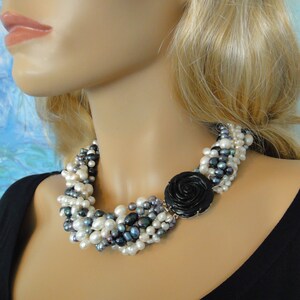 Puede incluir: Un collar de perlas de m&uacute;ltiples hilos con un colgante de rosa negra. El collar est&aacute; hecho de perlas blancas y grises y est&aacute; dispuesto en un patr&oacute;n en cascada. El colgante es una rosa negra grande con un acabado brillante.
