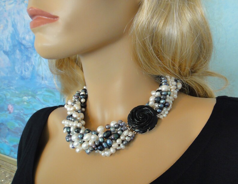 Puede incluir: Un collar de perlas de m&uacute;ltiples hilos con un colgante de rosa negra. El collar est&aacute; hecho de perlas blancas y grises y est&aacute; dispuesto en un patr&oacute;n en cascada. El colgante es una rosa negra grande con un acabado brillante.