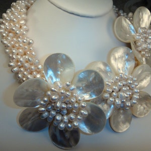 Puede incluir: Un collar de perlas blancas con un racimo de flores de concha blanca. El collar está hecho de pequeñas perlas redondas y las flores son grandes y delicadas.