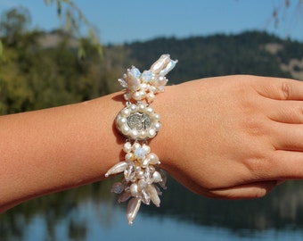 May include: A white pearl bracelet with a silver charm featuring a tree design. The bracelet is made with a variety of pearl shapes and sizes, including baroque pearls.