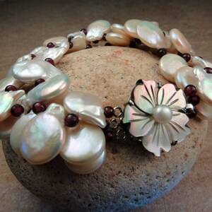 Puede incluir: Un brazalete de perlas blancas con un cierre en forma de flor. El brazalete está hecho de múltiples hileras de perlas y presenta una sola perla en el centro del cierre en forma de flor. Las perlas son iridiscentes y tienen un color ligeramente blanquecino. El brazalete está acentuado con pequeñas cuentas de color rojo oscuro.