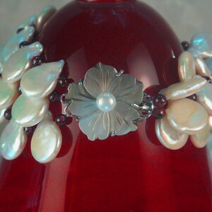 May include: A silver flower charm bracelet with a white pearl in the center of the flower. The bracelet is made with white and iridescent shell beads and burgundy beads.