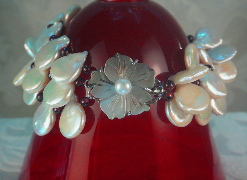 May include: A silver flower charm bracelet with a white pearl in the center of the flower. The bracelet is made with white and iridescent shell beads and burgundy beads.