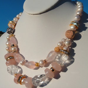 Puede incluir: Un collar de dos hileras con cuentas facetadas de color rosa y transparente, perlas blancas y detalles dorados y plateados.