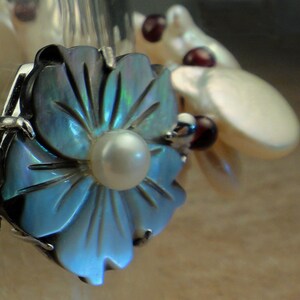 May include: A blue iridescent flower-shaped pendant with a white pearl in the center. The pendant is attached to a silver chain.