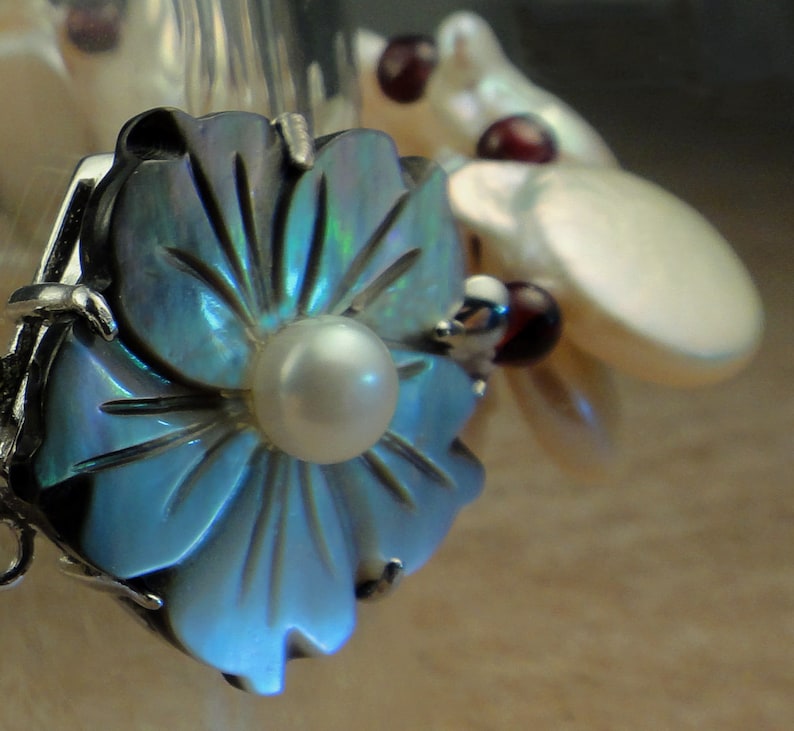 May include: A blue iridescent flower-shaped pendant with a white pearl in the center. The pendant is attached to a silver chain.