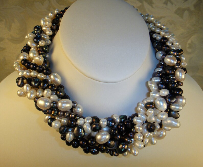 Puede incluir: Un collar de perlas de m&uacute;ltiples hilos con una mezcla de perlas blancas y negras. El collar est&aacute; hecho de una variedad de tama&ntilde;os y formas de perlas, creando un aspecto &uacute;nico y elegante.