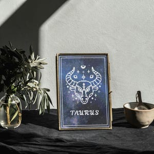 May include: A framed print of the Taurus zodiac sign, with a white bull illustration on a dark blue starry background. A small ceramic bowl with a spoon and a glass vase with greenery are also present.