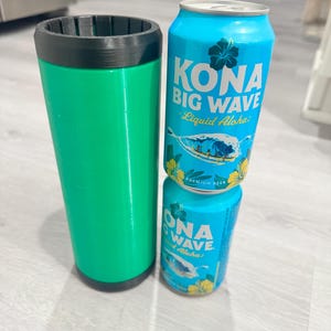 Puede incluir: Un soporte verde aislado para bebidas con borde negro, junto a dos latas de cerveza Kona Big Wave. Las latas son de color azul claro con texto blanco y un gráfico de olas. El soporte es cilíndrico.