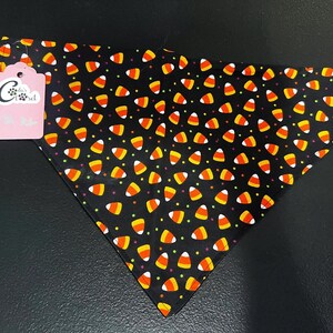 May include: Black triangular pet bandana patterned with candy corn in orange, yellow, and white. The bandana has a pink tag with the text "Cody's Closet" and a logo. The background is black.
