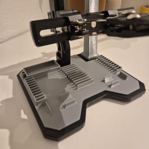Op de afbeelding: Een grijs en zwart 3D-geprint gereedschapshouder met meerdere compartimenten en een steunarm. De basis is grijs met een zwarte rand en de arm is zwart met zilveren accenten. Het ontwerp lijkt bedoeld om gereedschap te organiseren.