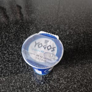 Könnte beinhalten: Ein Einzelportionsbecher Joghurt von YOGOS NATURE. Der Behälter ist blau und weiß, mit dem Markennamen und Produktinformationen auf dem Deckel. Der Deckel ist teilweise abgezogen und gibt den Blick auf den Joghurt frei.
