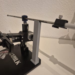 Könnte beinhalten: Ein schwarzes und silbernes Messerschärfsystem mit einem grauen Stützpfosten. Das System hat eine lange, silberne Stange und eine schwarze Basis mit dem Wort "KARILK" in silbernen Buchstaben. Zum Schärfen von Messern.