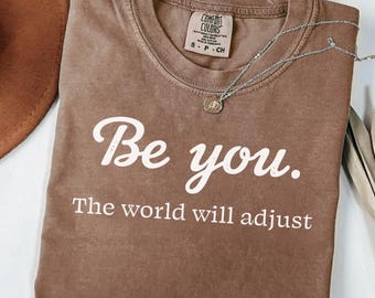 Puede incluir: Una camiseta marrón con las palabras "Be you. The world will adjust" en escritura blanca. Un collar plateado con un pequeño colgante descansa sobre la camiseta. Un sombrero marrón es parcialmente visible.