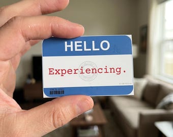 May include: A rectangular name tag sticker with a blue top section featuring the white text HELLO and a white bottom section with the red text Experiencing. A faint circular stamp is visible in the center, with a small barcode and text at the bottom.