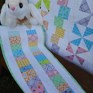 May include: A white, green, blue, and pink patchwork quilt with a white border. The quilt is made of squares and pinwheels. A white stuffed bunny rabbit is sitting on the quilt.