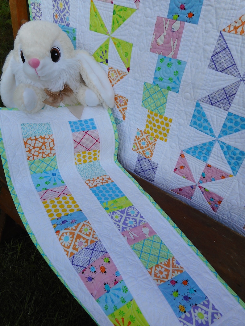 May include: A white, green, blue, and pink patchwork quilt with a white border. The quilt is made of squares and pinwheels. A white stuffed bunny rabbit is sitting on the quilt.