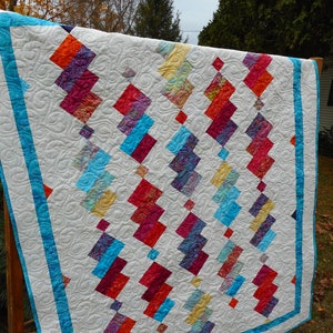 May include: A handmade quilt with a white background and a repeating pattern of colorful, angled blocks. The blocks feature vibrant hues of red, blue, orange, and purple. The quilt is bordered with a bright blue fabric and has intricate quilted stitching.