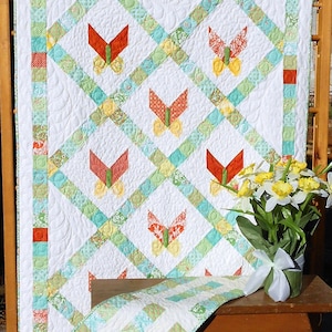 May include: A white quilt with a green, blue, and yellow patchwork design. The quilt features a repeating pattern of butterflies with orange, red, and yellow accents. The quilt is displayed on a wooden table with a bouquet of yellow daffodils in a vase.