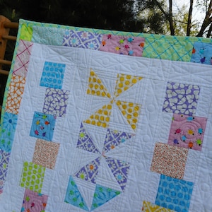 May include: A white quilt with a patchwork design featuring squares and triangles in various colors and patterns, including pink, yellow, blue, green, and purple. The quilt is quilted with a swirl design.