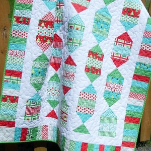 May include: A handmade Christmas quilt with a white background and a repeating pattern of colorful candy-shaped designs. The quilt features red, green, and turquoise fabrics with various patterns, including polka dots, stripes, and Christmas trees. The quilt is trimmed with a green border.