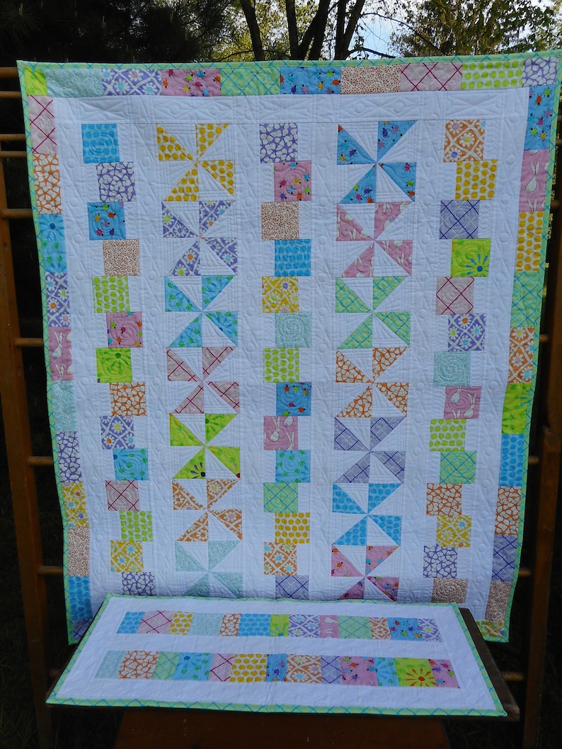 May include: A white quilt with a patchwork design featuring a variety of colorful fabrics. The quilt is made up of squares and triangles, and the fabric patterns include polka dots, flowers, and geometric shapes. The quilt is bordered with a light green trim.