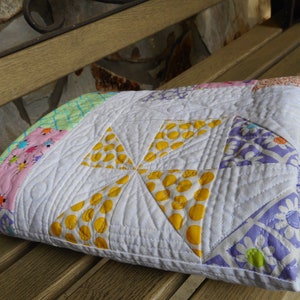 May include: A white quilt with a pinwheel design in yellow, pink, green, and purple fabrics. The quilt is folded and sitting on a wooden bench.