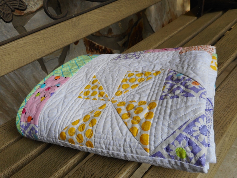 May include: A white quilt with a pinwheel design in yellow, pink, green, and purple fabrics. The quilt is folded and sitting on a wooden bench.