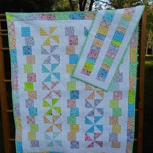 May include: A white quilt with a green, yellow, pink, and blue patchwork design. The quilt is made up of squares and triangles. It is hanging on a wooden rack.