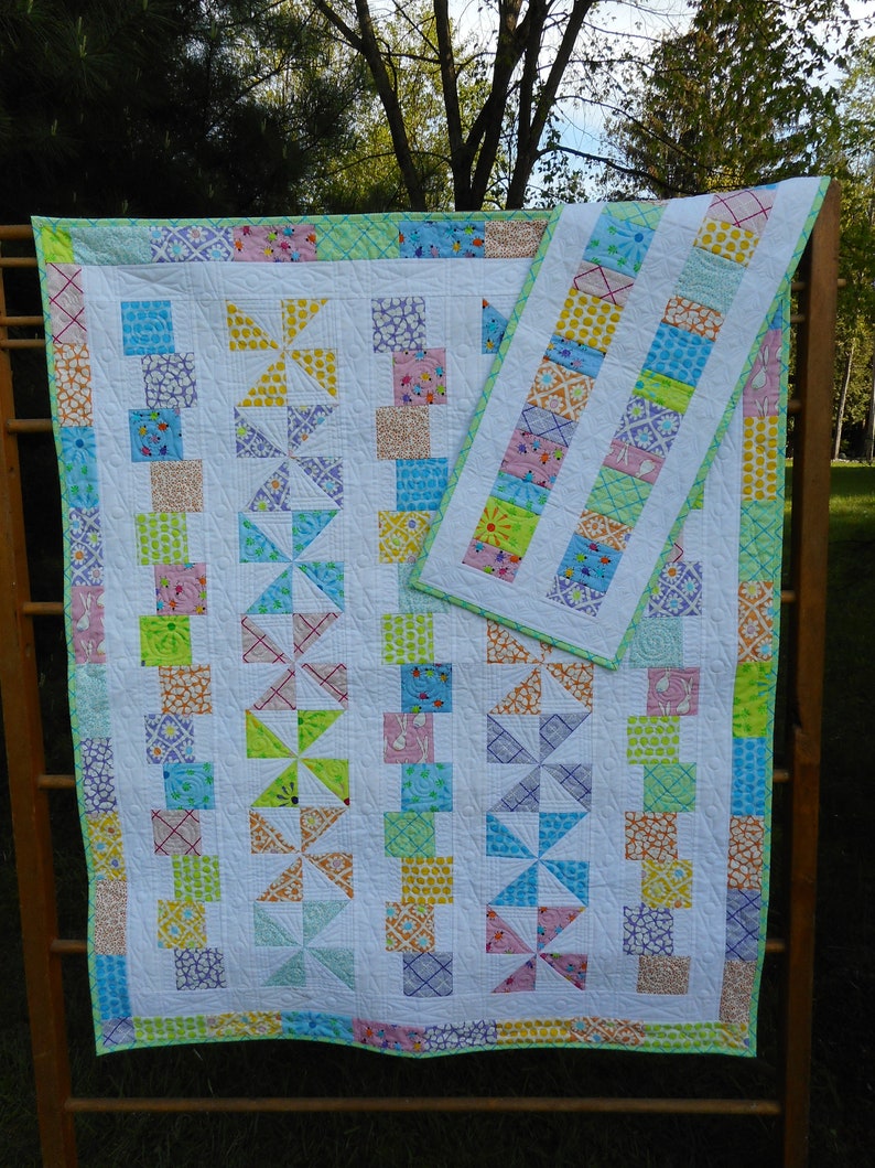 May include: A white quilt with a green, yellow, pink, and blue patchwork design. The quilt is made up of squares and triangles. It is hanging on a wooden rack.