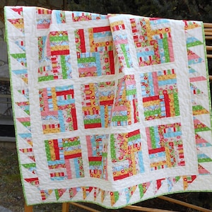 May include: A white quilt with a log cabin pattern in shades of red, green, yellow, blue, and pink. The quilt has a white border and a green and white border with a triangle pattern.