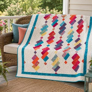 May include: A colorful quilt with a white background and a geometric pattern of stacked, angled rectangles in shades of red, orange, yellow, and blue. The quilt has a teal border and is draped over a wicker chair.