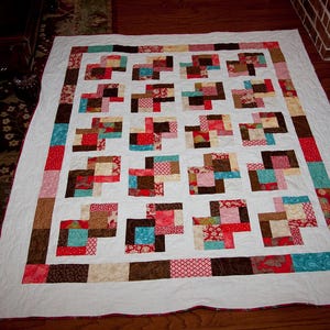 May include: A white quilt with a patchwork design featuring squares of red, brown, blue, and cream fabric. The quilt is made up of a grid of squares with a white background.