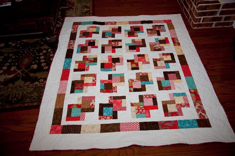 May include: A white quilt with a patchwork design featuring squares of red, brown, blue, and cream fabric. The quilt is made up of a grid of squares with a white background.