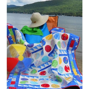 May include: A white and blue quilt with a beach theme. The quilt features a variety of colorful circles and a beach ball. The quilt is folded and sitting on a sandy beach. The text "Beach Blanket Bingo" is written below the quilt. The text "Fabrics from "Ocean View" by Paul Brent for Moda" is written below the title.