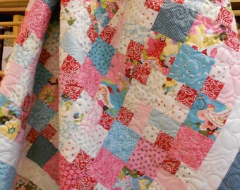 May include: A patchwork quilt with a variety of pink, blue, and white fabrics. The quilt is made up of squares of different patterns and colors, and it is quilted with a simple diamond pattern.