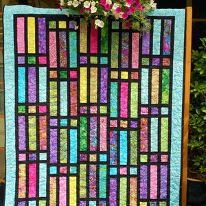 May include: A colourful quilt with a stained glass window design. The quilt is made up of various shades of pink, purple, blue, green, and yellow fabric. The quilt is framed with a black border and a light blue border.