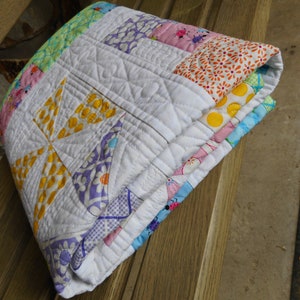 May include: A white quilt with a patchwork design featuring yellow, pink, blue, and green floral patterns. The quilt is folded and sitting on a wooden surface.