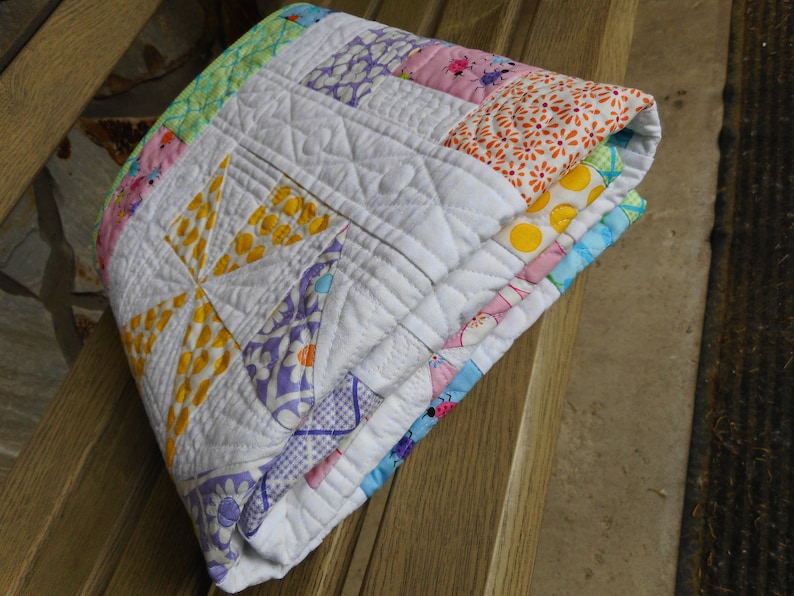 May include: A white quilt with a patchwork design featuring yellow, pink, blue, and green floral patterns. The quilt is folded and sitting on a wooden surface.